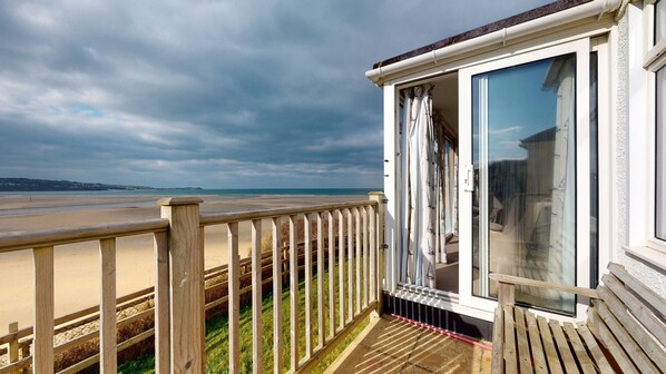Une terrasse en bois avec une porte vitrée donnant sur une pièce offrant une vue sur la plage et l’océan.