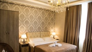 A hotel room with a large bed, two bedside lamps, a chandelier, a wooden wardrobe, and a small table with a glass of water and a bowl of fruit. - De'Mar Hotel (Bishkek)
