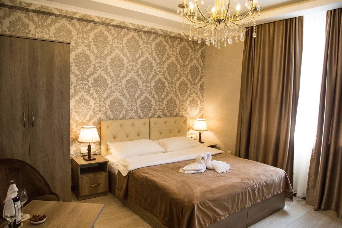A hotel room with a large bed, two bedside lamps, a chandelier, a wooden wardrobe, and a small table with a glass of water and a bowl of fruit.