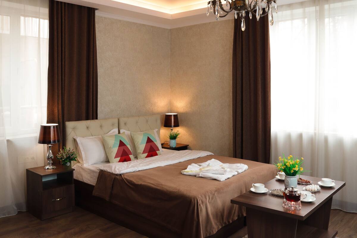 A hotel room with a bed, bedside tables, a desk with a tea set, and a window with curtains.