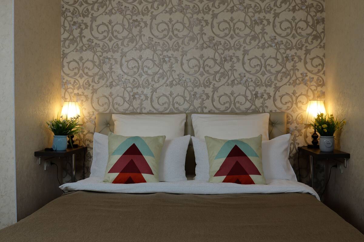 A neatly made bed with a patterned headboard, two bedside tables with lamps and potted plants, and a decorative pillow.