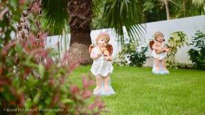 Two angel figurines on a grassy lawn with palm trees and a white wall in the background.