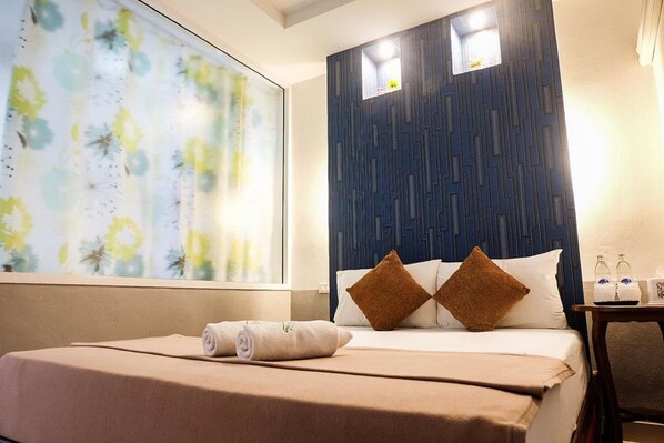 Habitación de hotel con cama, un ventanal con vistas a flores, una pared oscura con diseños geométricos y una mesita con artículos de aseo.