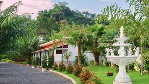 A white building with a red roof, surrounded by lush greenery and a white fountain.