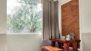 A corner room with a window, a wooden bench, a small table with bottles, and a brick wall.