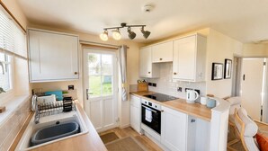 A compact kitchen with a sink, stove, and countertop, featuring white cabinetry and a small dining area. - G03 Lanteglos (G3), Riviere Towans (Hayle)