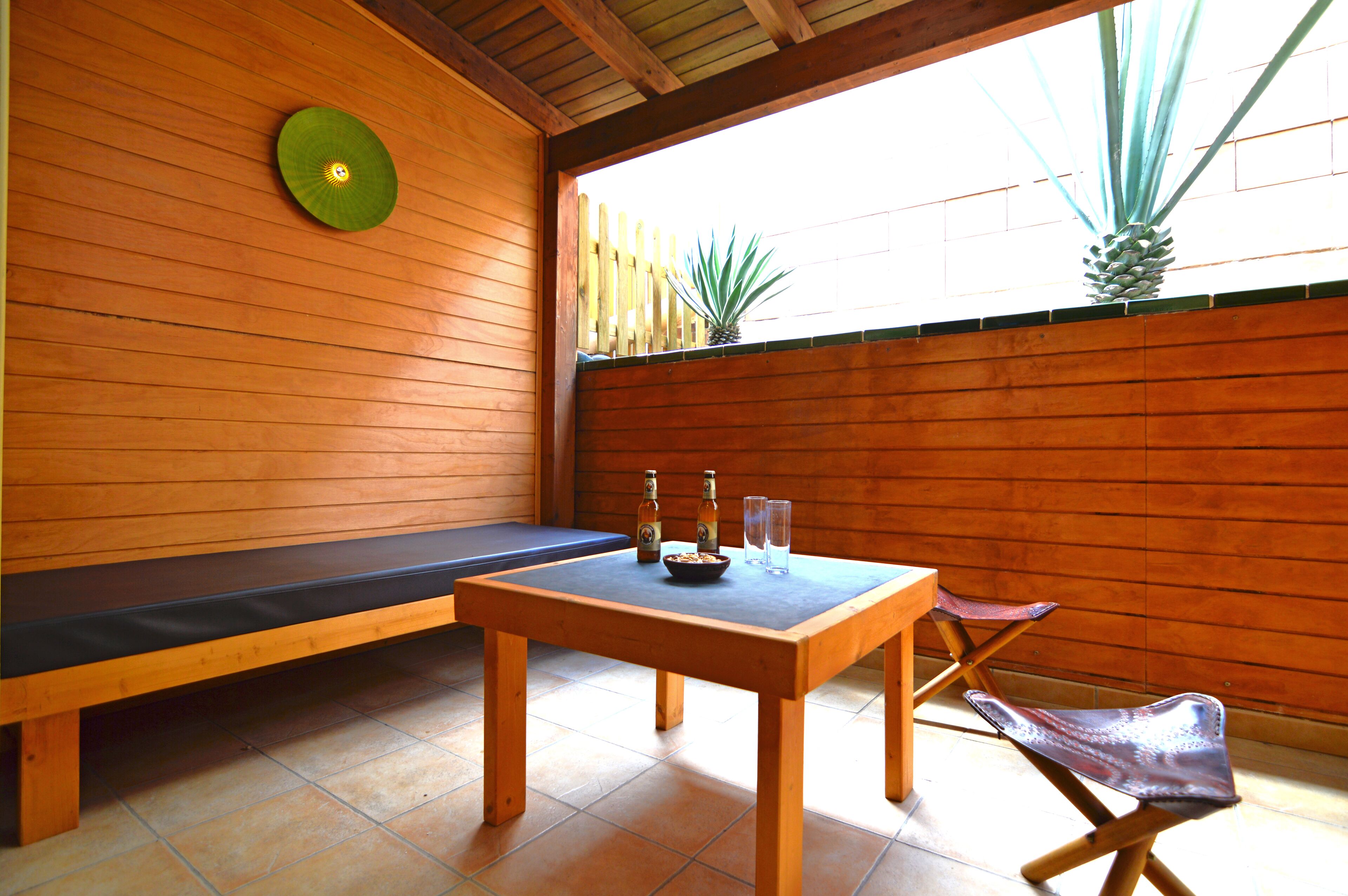 A wooden interior with a bench, a table with bottles and glasses, and a view of a wooden fence and plants outside.