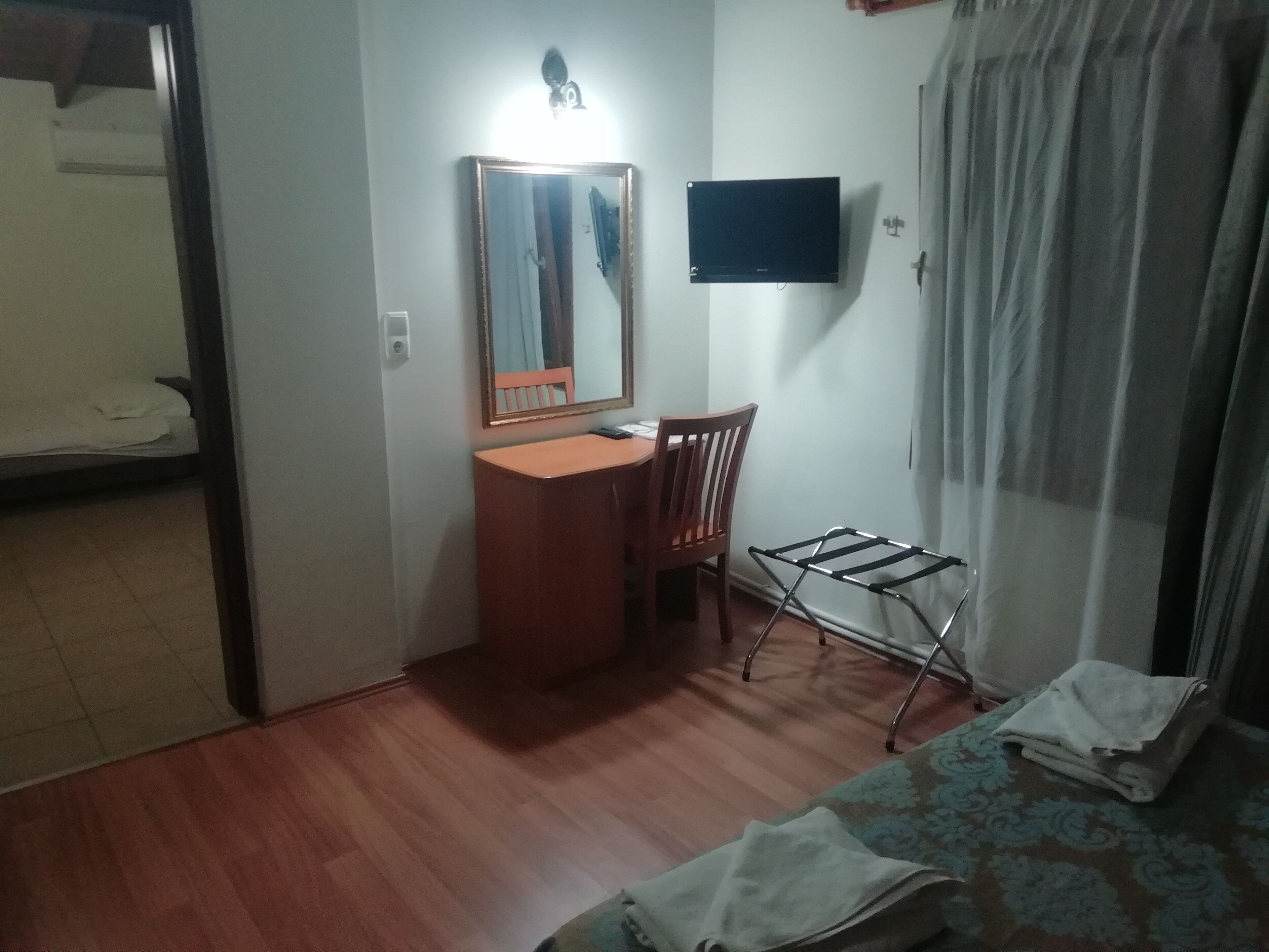 A small, dimly lit room with a wooden desk, a chair, a mirror, a television mounted on the wall, and a bed with a patterned bedspread.
