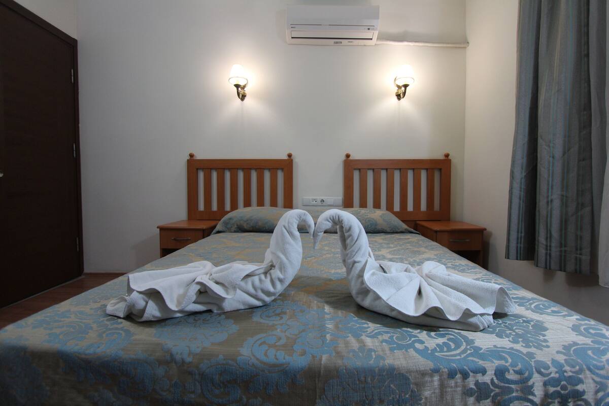A hotel room with two beds, a wooden headboard, a nightstand, a wall-mounted air conditioner, and two wall sconces.