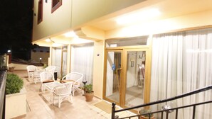 Un patio de hotel con mobiliario de mimbre blanco, plantas en macetas y una puerta de cristal que da a un espacio interior.
