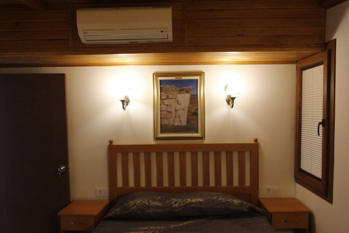 A bedroom with a wooden ceiling, a bed with a patterned bedspread, two bedside tables, a wall-mounted air conditioner, and a framed picture above the bed.