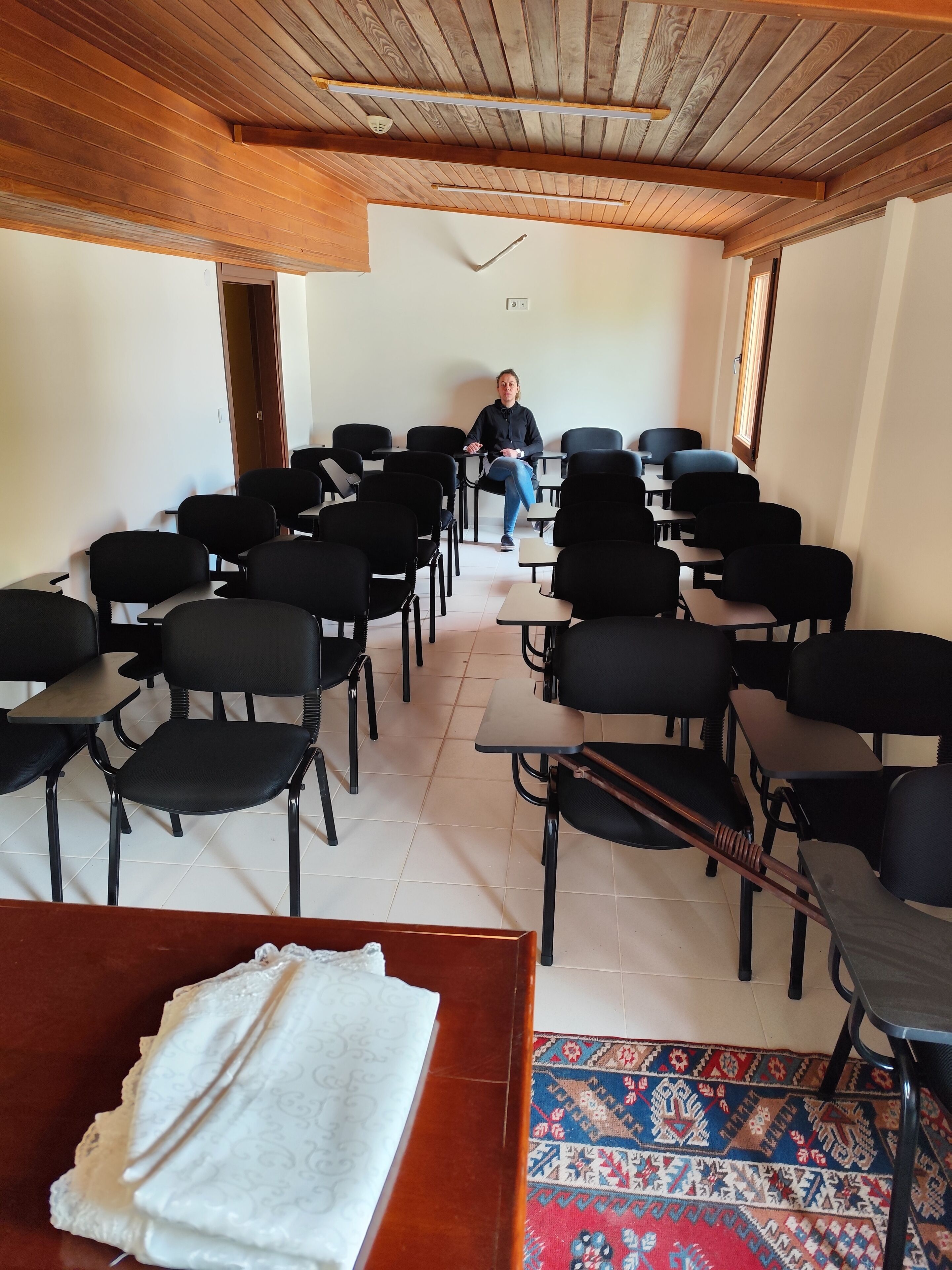 A conference room with rows of black chairs and tables, a wooden ceiling, and a patterned rug on the floor.