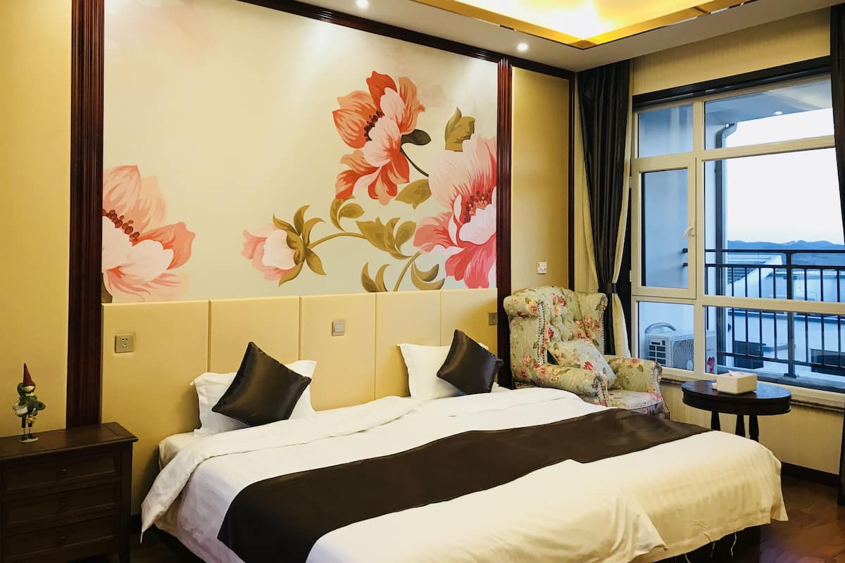 Une chambre d’hôtel avec un grand lit, une fresque murale à motifs floraux, une fenêtre donnant sur un beau paysage et une petite table sur laquelle se trouve une boîte.