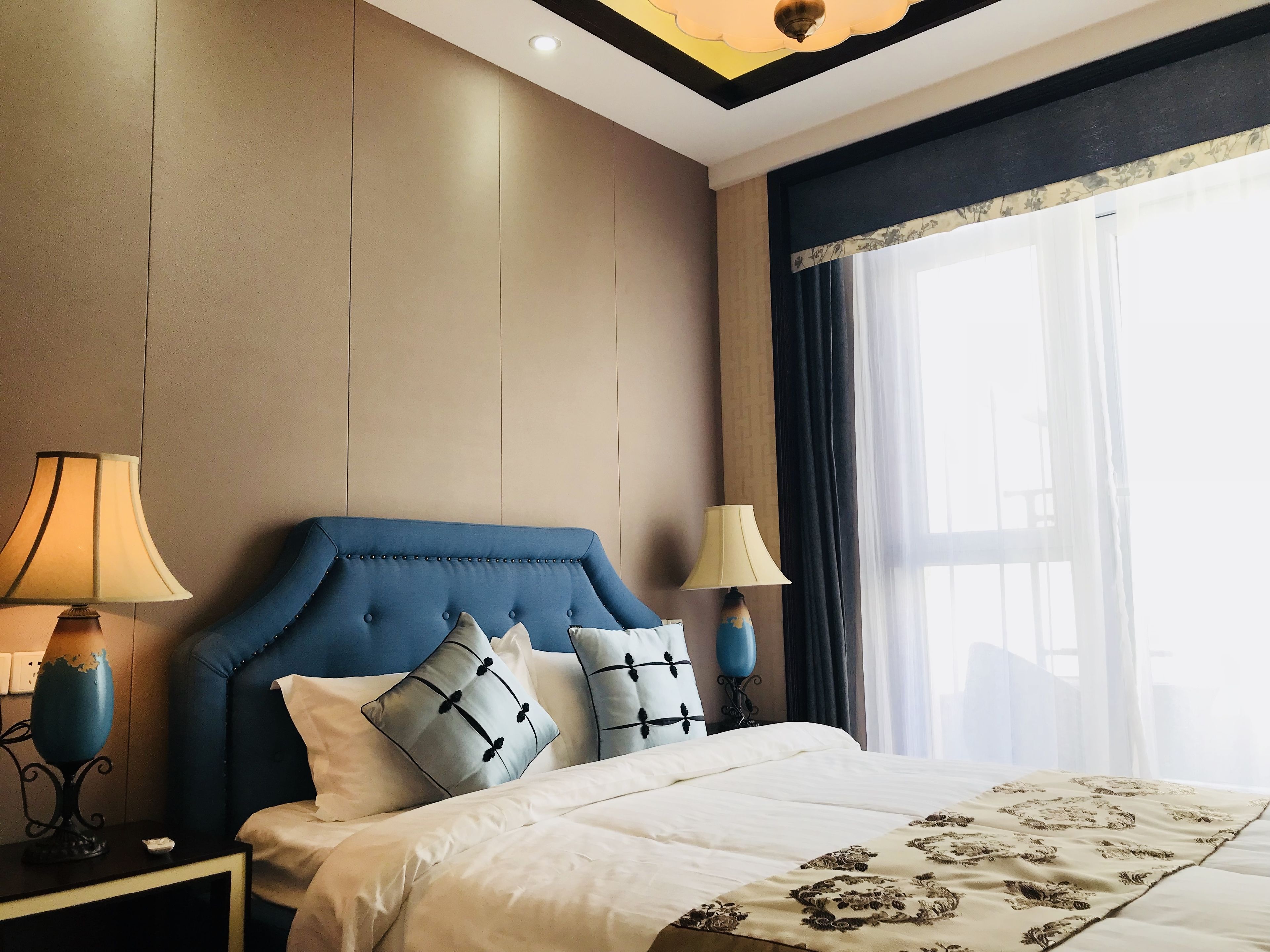 Quarto de hotel com uma cama grande, duas luminárias de cabeceira, uma janela com cortinas e um lustre.