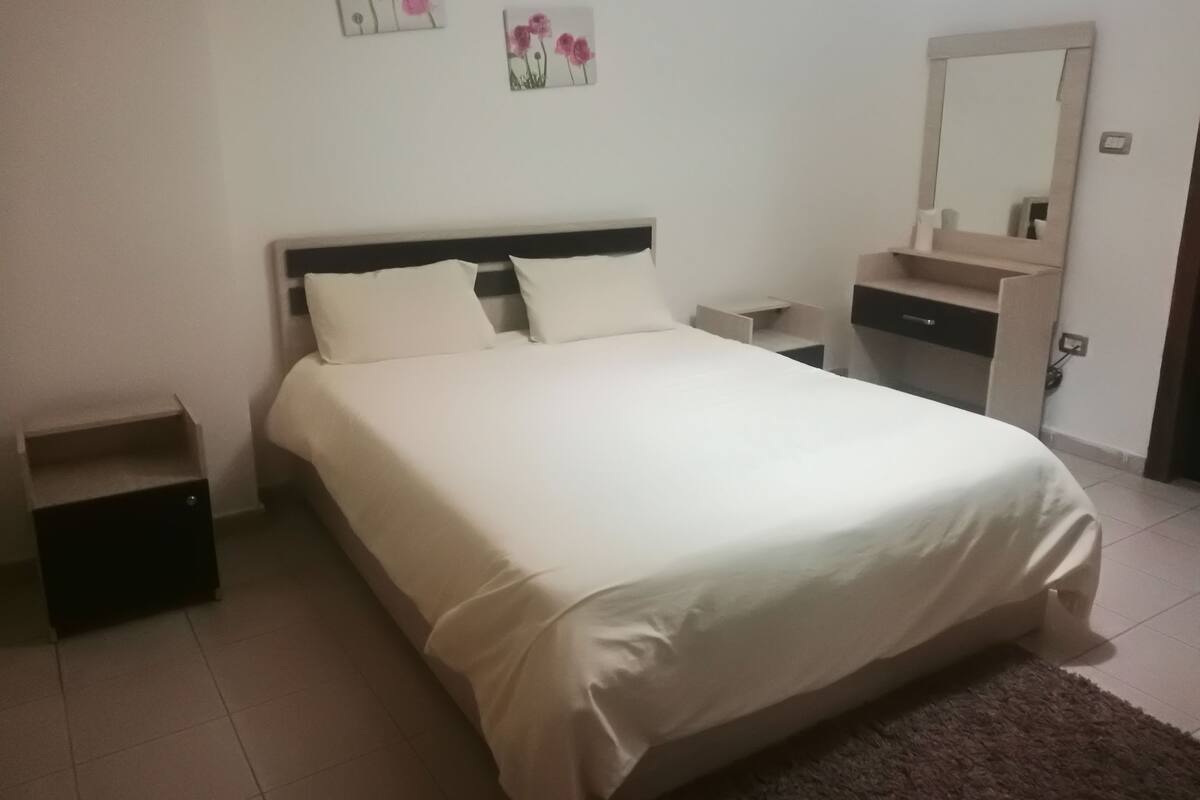 Een netjes opgemaakt bed met witte beddengoed, een nachtkastje, een spiegel en een klein plankje.