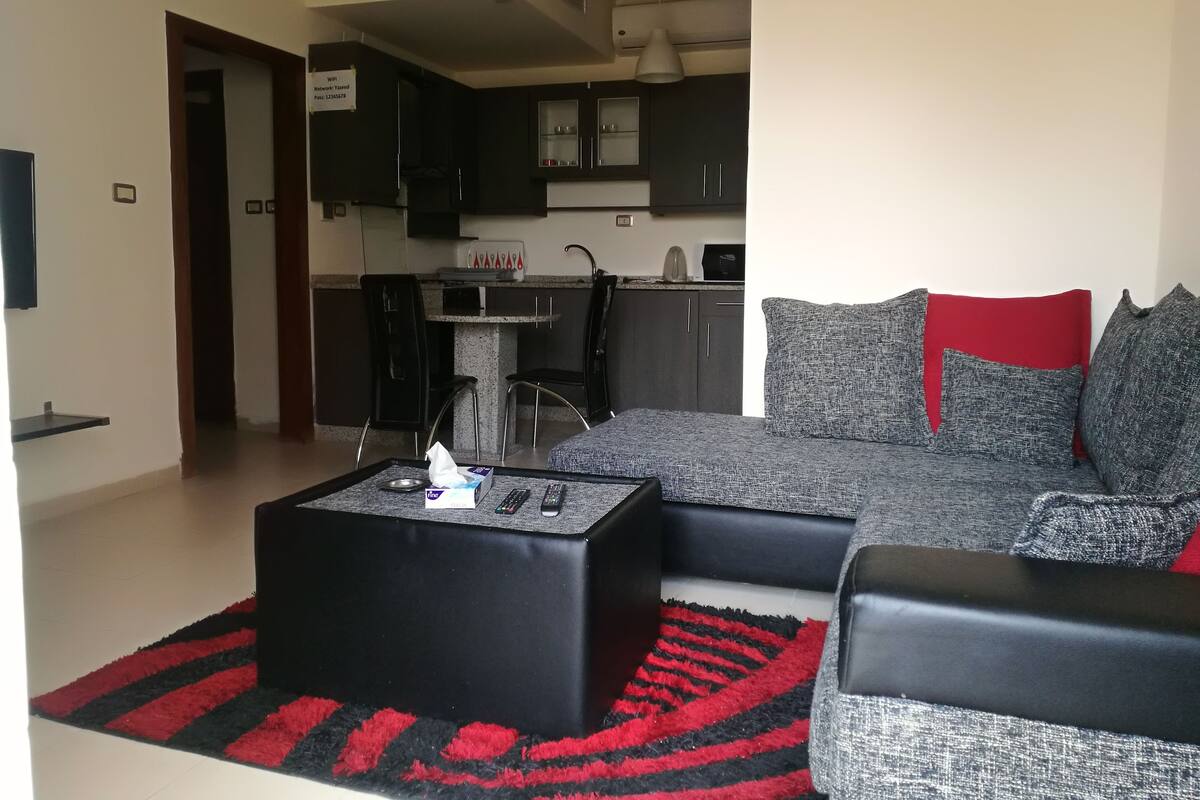 Een woonkamer met een zwarte salontafel, een grijze bank en rode kussens. Achtergrond is een keuken met donkere kastjes en een wit aanrecht.