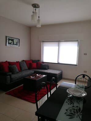 A living room with a grey sofa, red and grey cushions, a coffee table, a dining table with chairs, and a window with blinds.
