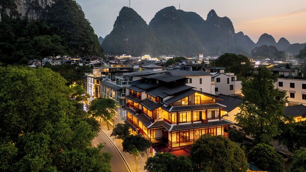 A traditional building with multiple stories and a modern structure nearby, surrounded by lush greenery and mountains in the background. - Yangshuo Riverview Hotel (Guilin)