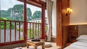 A hotel room with a wooden floor, a bed with white bedding, a small wooden table with a tray, a wicker ottoman, and a view of a mountain through large windows. - Yangshuo Riverview Hotel (Guilin)