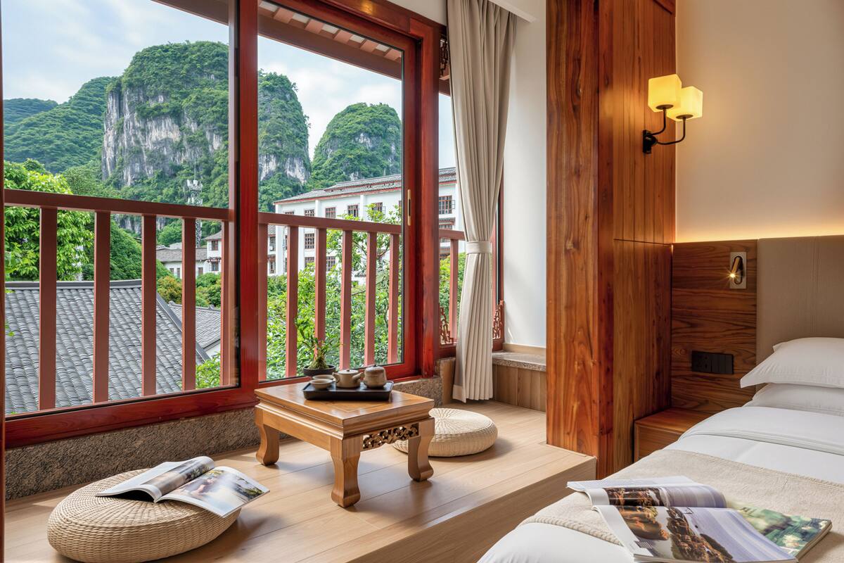 A hotel room with a wooden floor, a bed with white bedding, a small wooden table with a tray, a wicker ottoman, and a view of a mountain through large windows.
