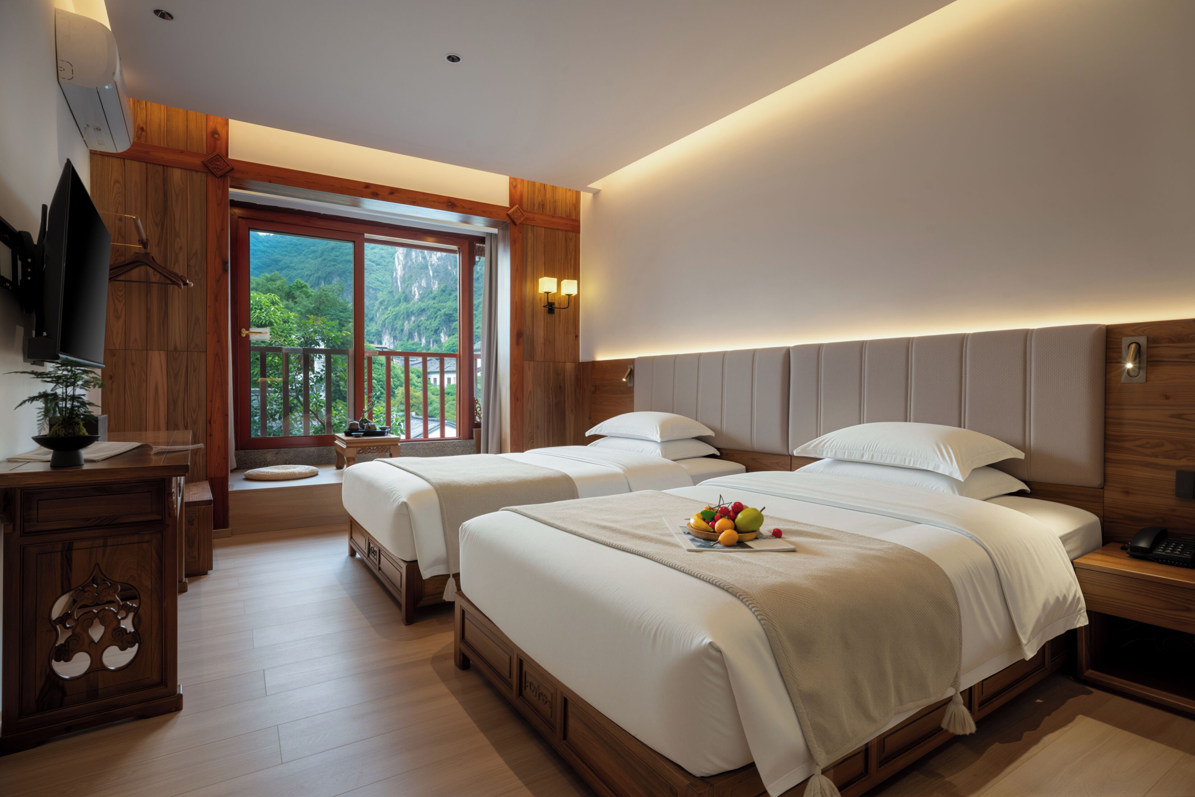 A hotel room with two beds, a wooden dresser, a TV, and a balcony with a view of greenery.