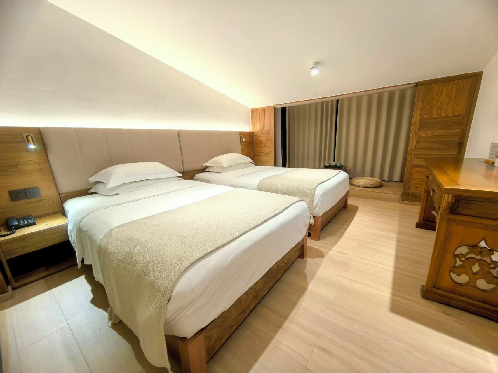 Habitación de hotel con dos camas, una mesita de noche de madera con un teléfono, un escritorio con una lámpara y un ventanal grande con cortinas.