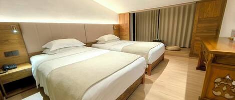 Habitación de hotel con dos camas, una mesita de noche de madera con un teléfono, un escritorio con una lámpara y un ventanal grande con cortinas.