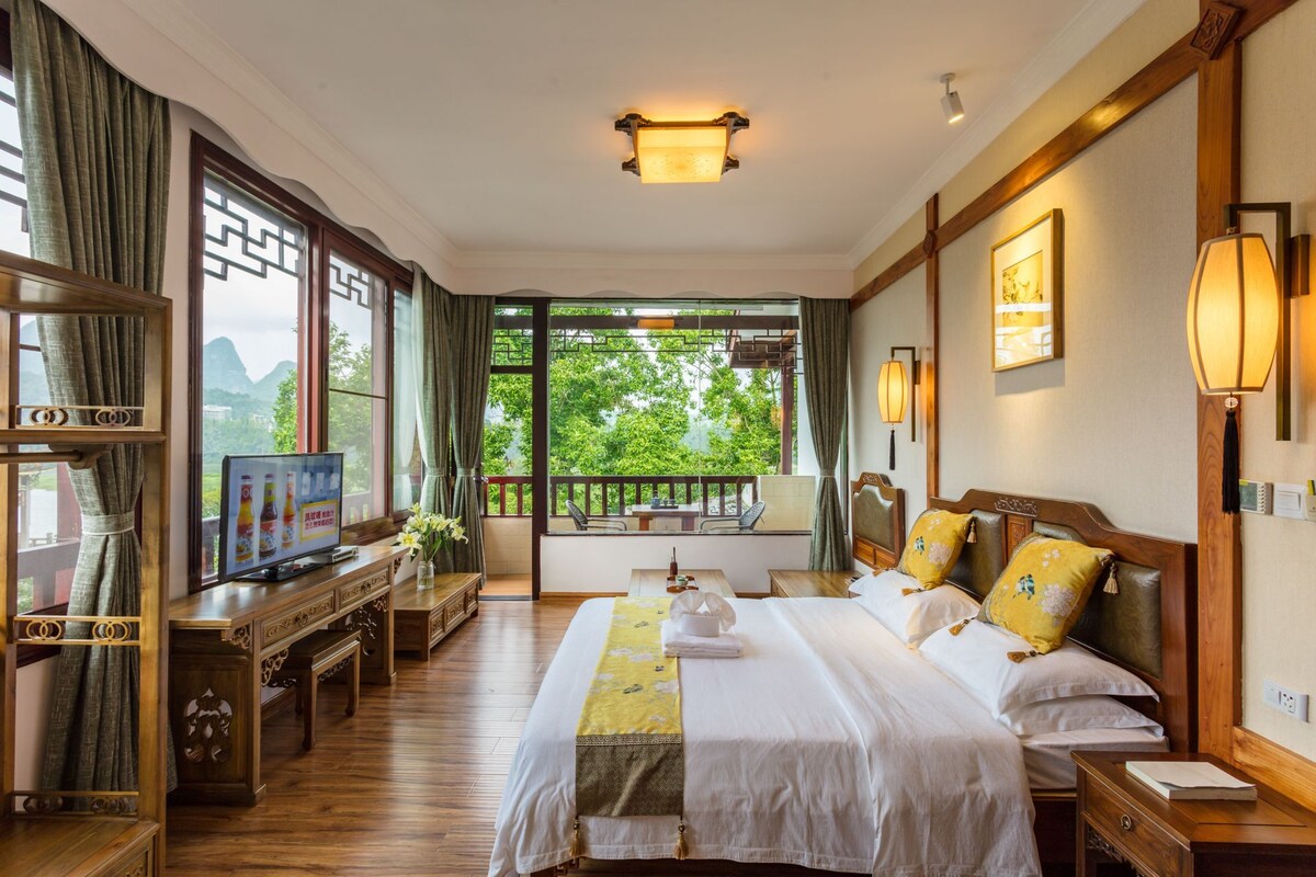 A hotel room with a large bed, wooden furniture, a TV, and a view of greenery.
