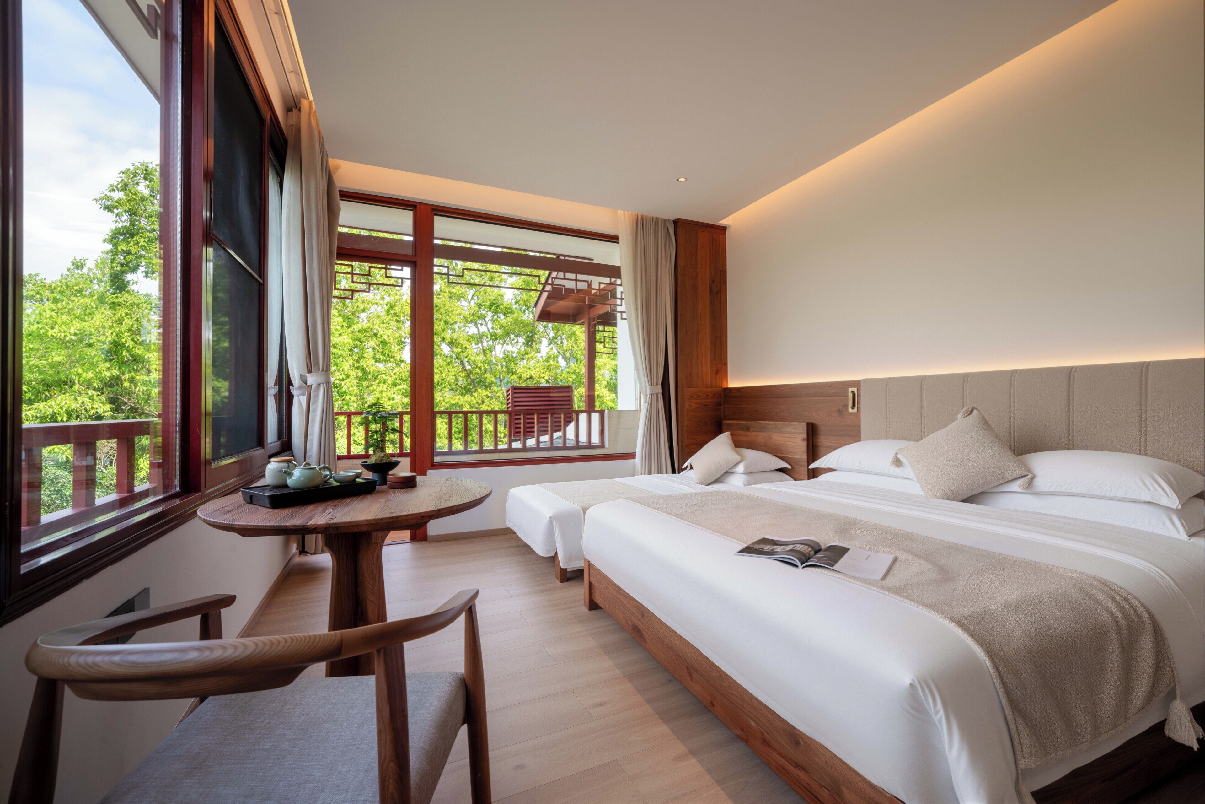 A hotel room with a large bed, a small table, a chair, and a balcony with a view of trees.
