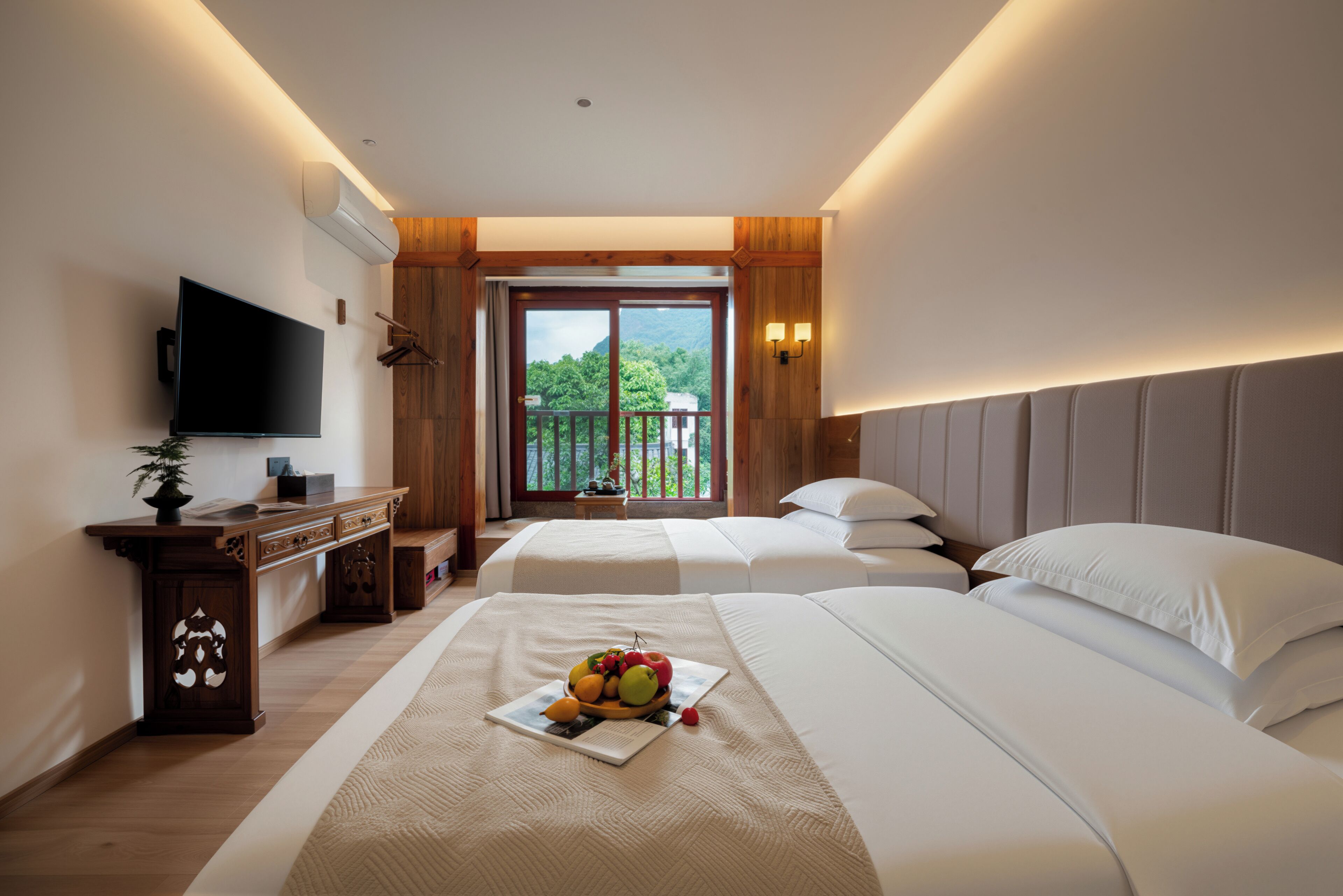 A hotel room with two beds, a flat-screen TV, a wooden desk, and a balcony with a view of greenery.