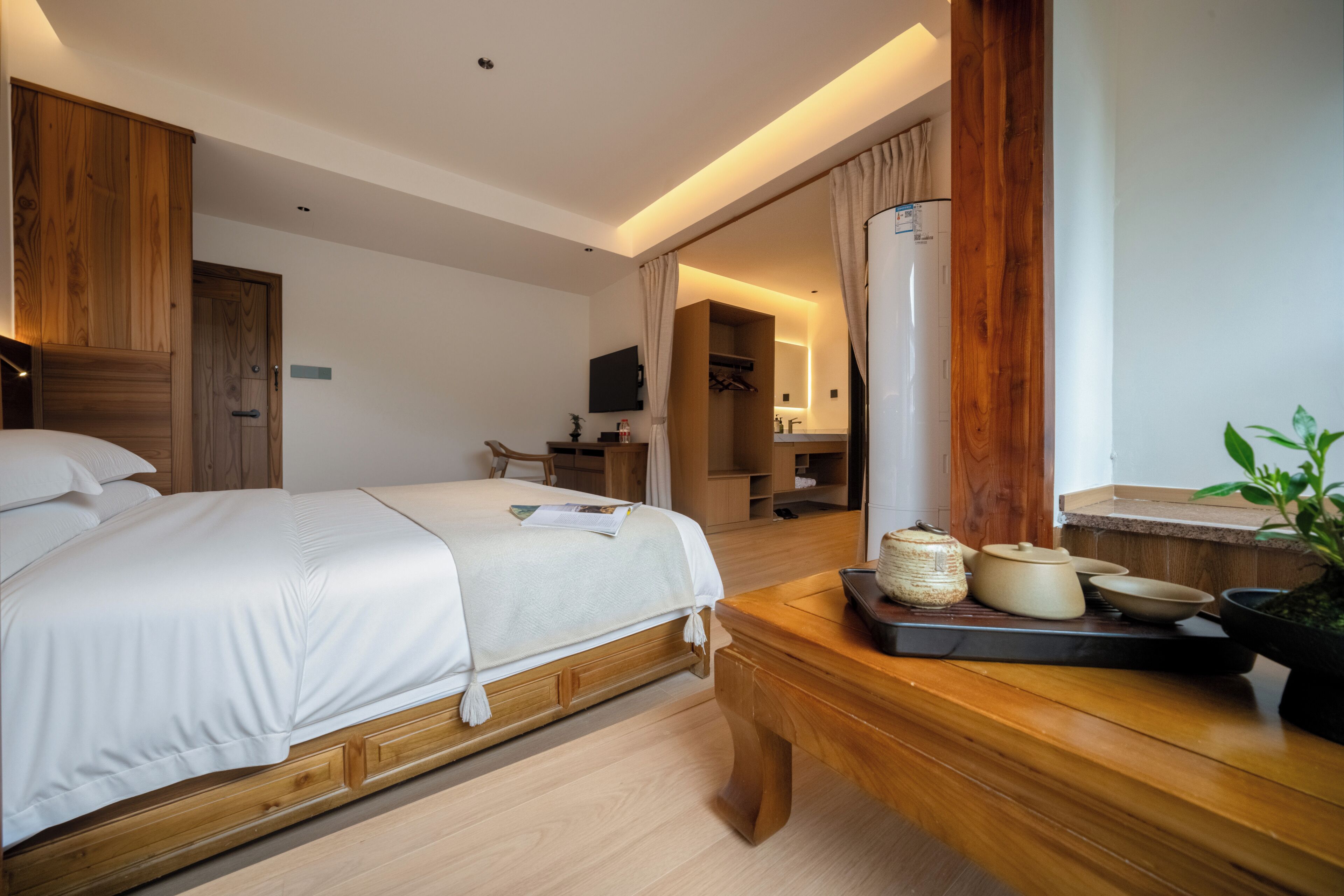 A modern hotel room with a large bed, wooden furniture, a desk, a television, and a bathroom visible through an open door.