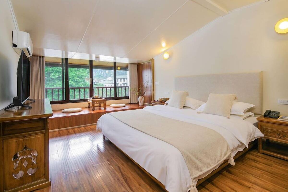 A bedroom with a large bed, wooden flooring, a TV on a wooden cabinet, and a balcony with a view of greenery.