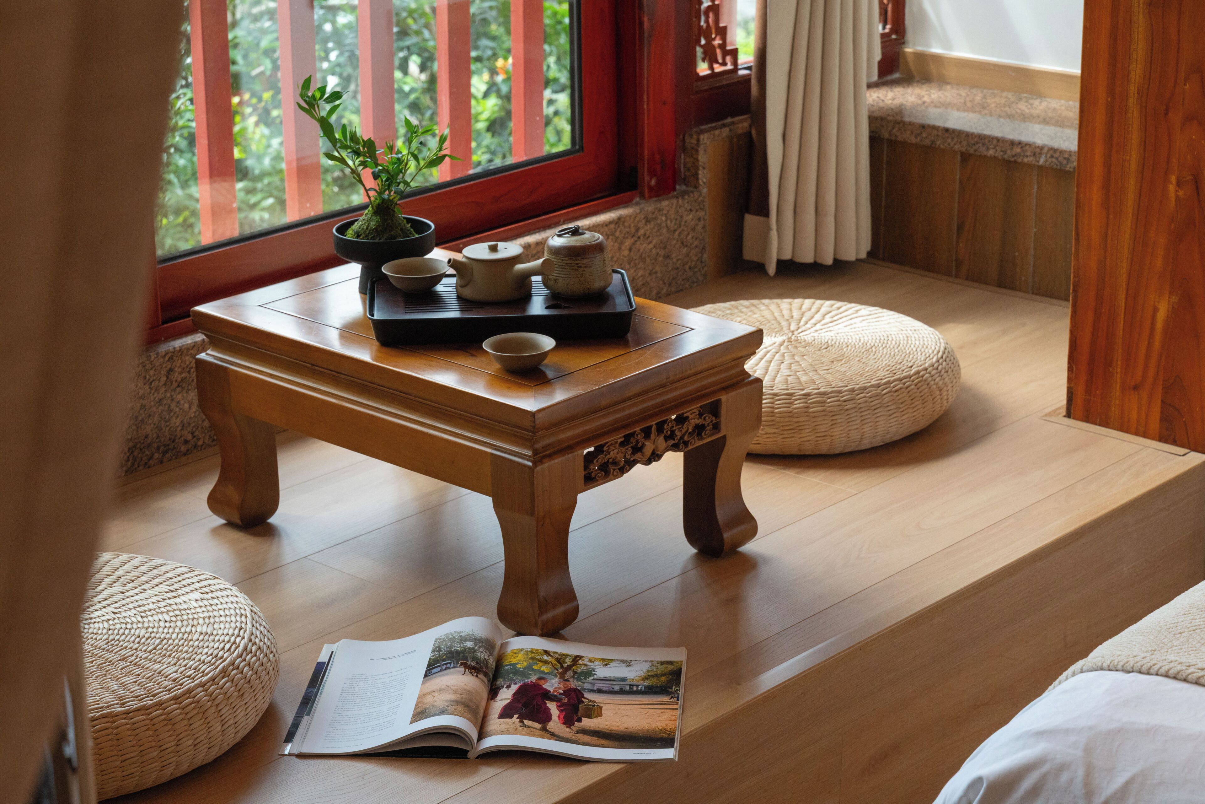 A wooden table with a tray of tea sets and a potted plant, a magazine on the floor, and a view of greenery through a window.
