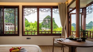 A room with a bed, a table with a teapot, a chair, and a view of mountains through large windows. - Yangshuo Riverview Hotel (Guilin)