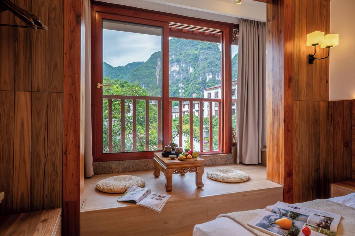 A room with wooden walls, a large window with a view of mountains, a small table with fruit, and a bed with a magazine.