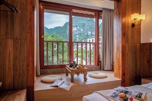 Ein Zimmer mit Holzwänden, einem großen Fenster mit Bergblick, einem kleinen Tisch mit Obst und einem Bett mit einer Zeitschrift.