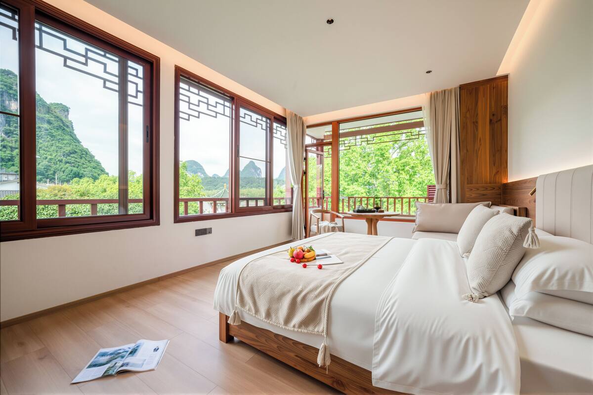 A bedroom with a large bed, a view of greenery, and a dining area with a table and chairs.