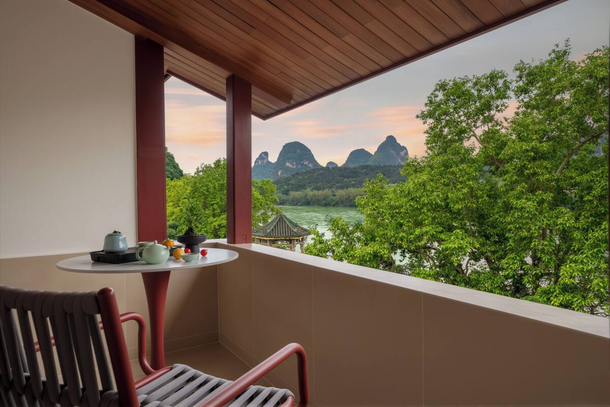 A balcony with a view of mountains and a river, a table with a teapot and cups, and a chair.