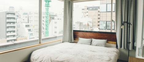 A hotel room with a large bed, a wooden headboard, a window with curtains, and a view of the cityscape.