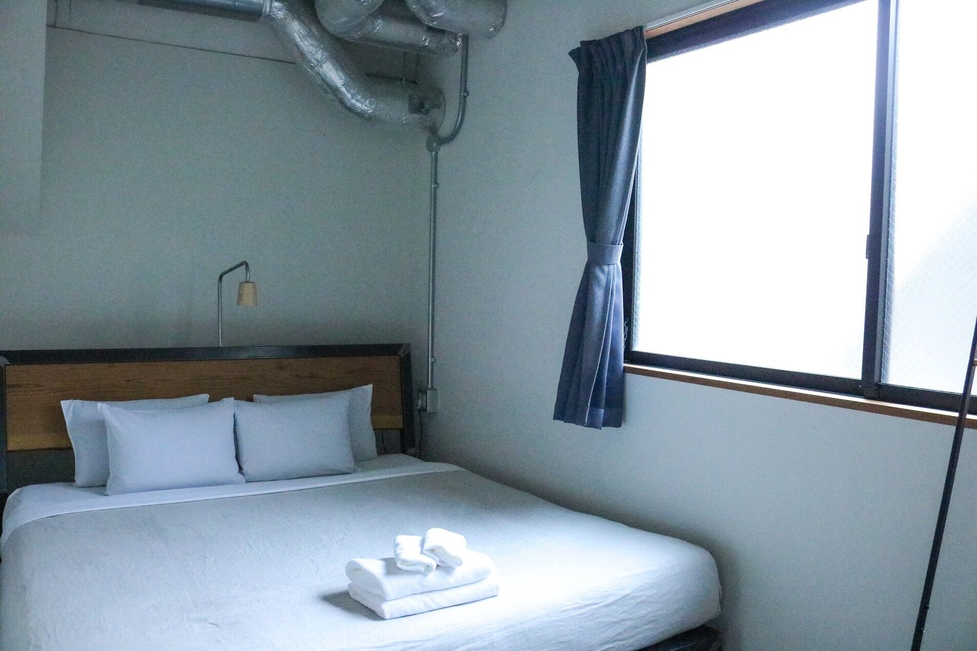 A hotel room with a bed, white bedding, a neatly folded towel, a wooden headboard, a window with curtains, and a wall-mounted lamp.