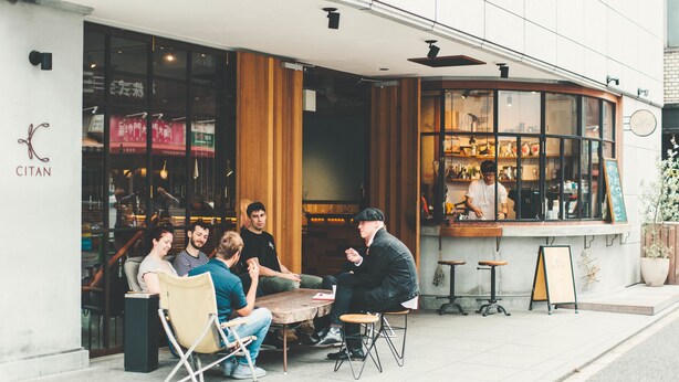 Een groep mensen zit aan een buitentafel voor een café met een houten interieur en grote ramen.