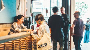 Een groep mensen bij een balie, mogelijk een café of winkel, met een bord waarop 'CLSA' staat.