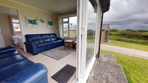 A cozy living room with a blue sofa, a small table, and a window overlooking a grassy area. - D58 Penlee, Riviere Towans (Hayle)