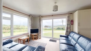 A living room with a sofa, a television on a stand, and a view of the outdoors through large windows. - D58 Penlee, Riviere Towans (Hayle)