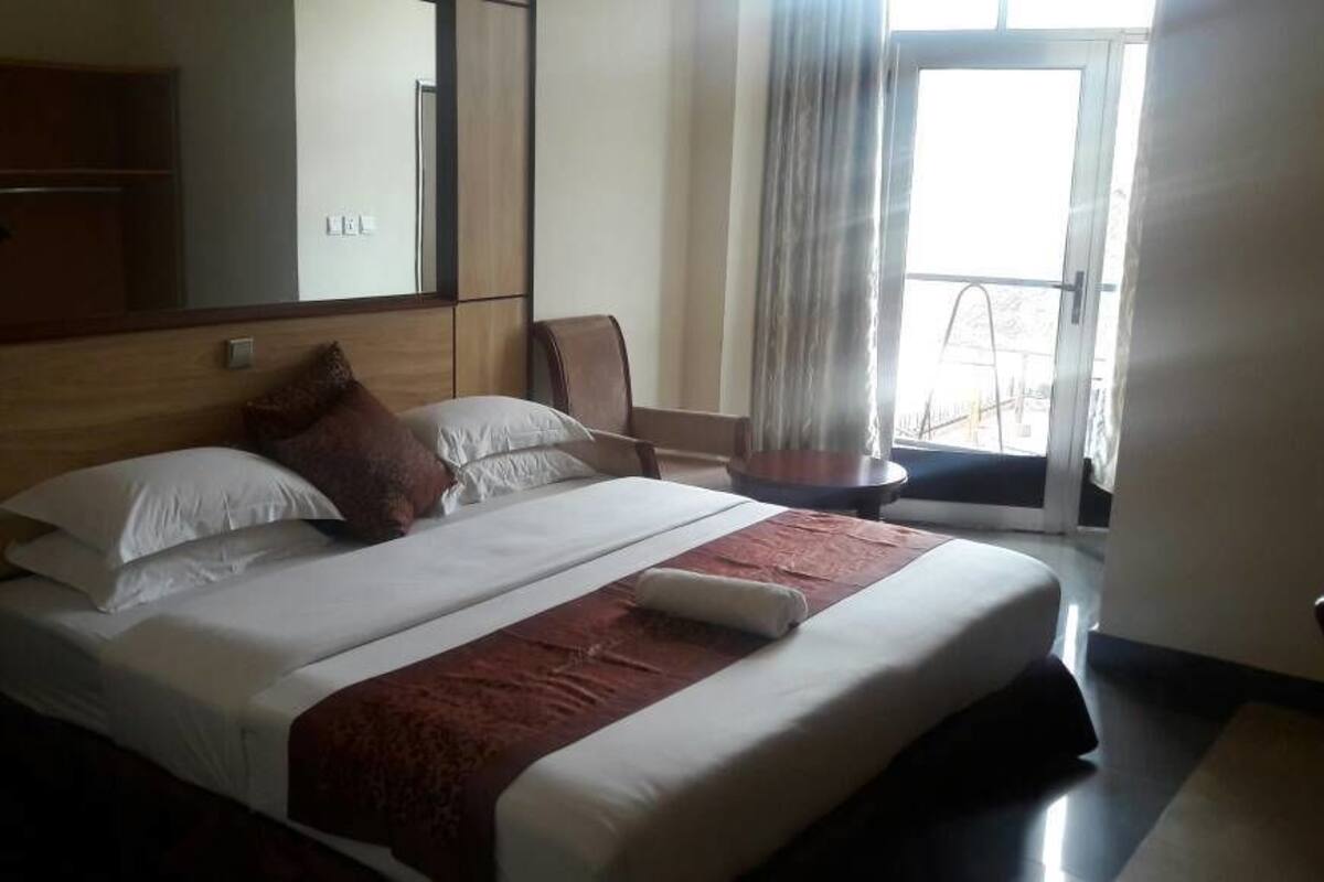 A hotel room with a large bed, a wooden headboard, a mirror, a window with curtains, and a small table.