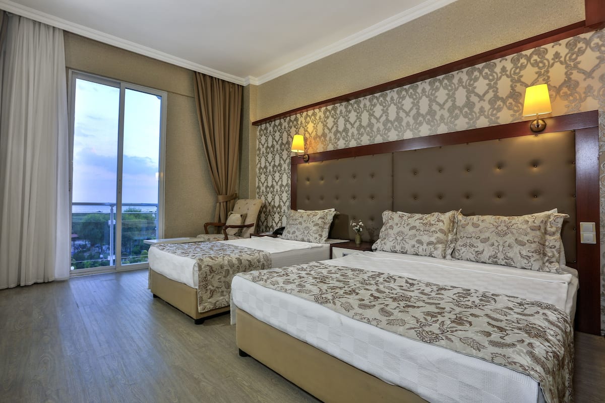 A hotel room with two beds, a large window with curtains, a headboard with a light fixture, and a view of the outdoors.