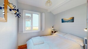 A bedroom with a large bed, a window with blinds, a bedside table with a lamp, and a painting on the wall. - C23 Wilmar, Riviere Towans (Hayle)