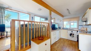 A compact kitchen with a washing machine, a dining area with a table and chairs, and a balcony with a view of the sea. - C23 Wilmar, Riviere Towans (Hayle)