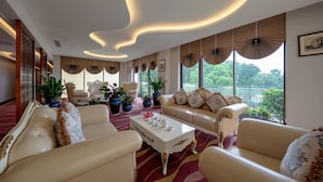 A spacious lounge with a white sofa set, a coffee table, and large windows offering a view of greenery. - Phoenix Resort Bac Ninh (Vu Ninh)