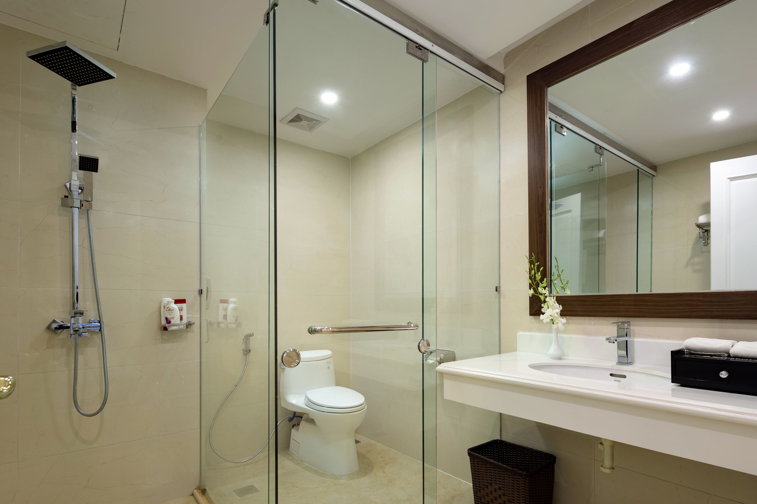 Un baño moderno con ducha envuelta en vidrio, inodoro blanco, lavamanos con espejo y un cubo de basura.